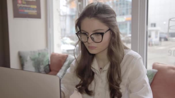 Hermosa chica caucásica en los vidrios en un café con el portátil — Vídeo de stock