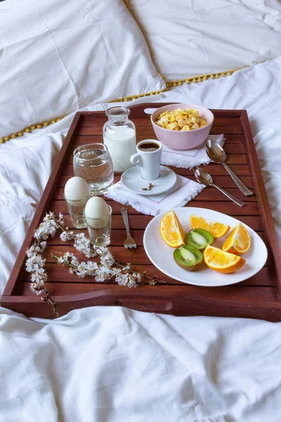 Romantic breakfast with flower branch in bed on wooden tray with corn flakes, fruits, milk, eggs and coffe. Window light, copy space