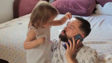 Sakallı baba akıllı telefonuyla uzaktan kumandayla çalışıyor. Küçük kızı yüzüne parlak çıkartmalar yapıştırıyor. Ona sarılıp öpüyor..