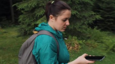 Sırt çantalı ve yağmurluk giyen bir kadın haritayı açmak için Smart Phone kullanıyor, ormanda tek başına yürüyor..