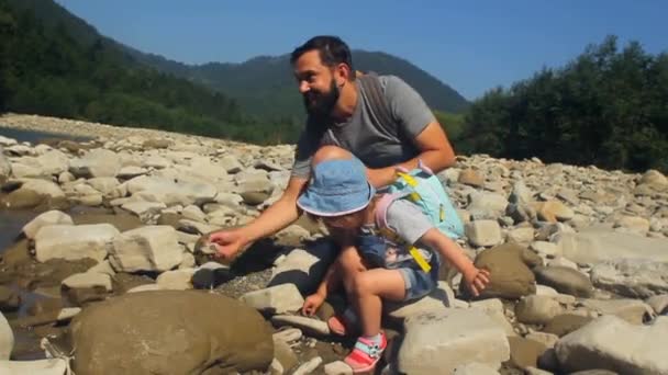 Tata z córeczką wrzuca kamienie do górskiej rzeki. Podróżowanie z dziećmi — Wideo stockowe