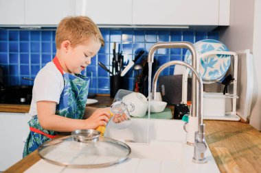 Önlüklü bir çocuk evde bulaşık yıkıyor.