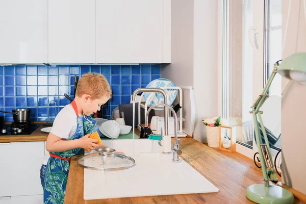 Un niño en un delantal lava los platos en la cocina en casa Fotos De Stock Sin Royalties Gratis