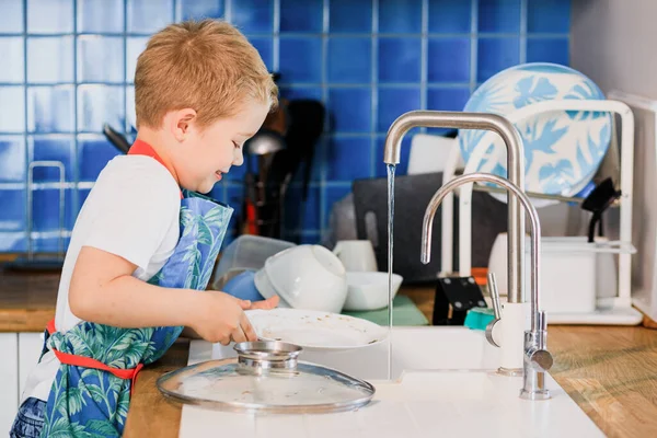 Un niño en un delantal lava los platos en la cocina en casa Fotos De Stock