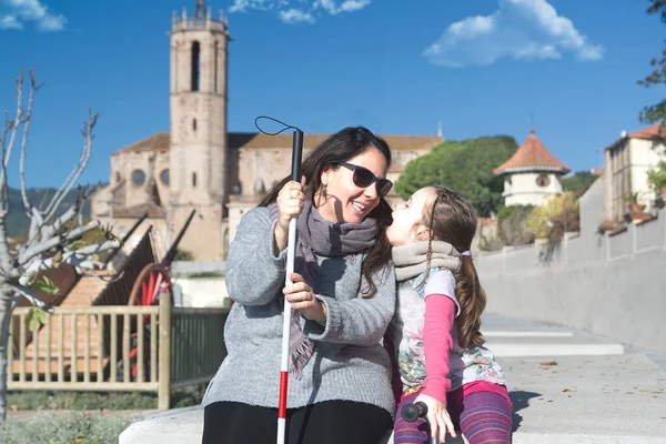 Blind mother with a white cane and her daughter talking sitting on a bench in the park and smiling. Visually impaired lifestyle concept with empty copy space.