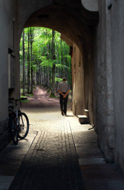 Yaşlı bir adam Trento, İtalya 'nın merkezindeki dar bir sokaktan, dalların arasından süzülen güneş ışığıyla huzurlu bir bahçeye doğru yürür.