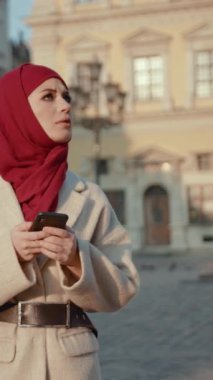 Canlı kırmızı bir başörtüsü içinde şık bir kadın, elinde bir telefonla, düşünceli bir şekilde uzaktan bakıyor.