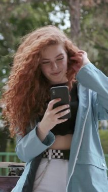 Canlı ve kıvırcık saçlı modaya uygun genç bir kadın lüks bir park ortamında akıllı telefonuna tamamen dalmış durumda.