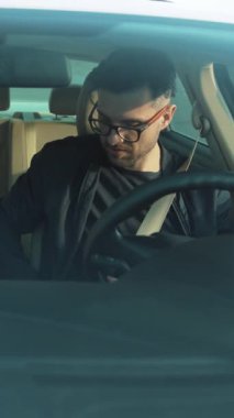 Moda gözlük takan şık bir genç adam rahat bir şekilde arabada oturuyor, kendine güveni ve modern titreşimleri yayıyor.