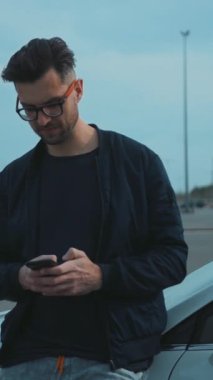 Havalı bir adam bulutlu bir gökyüzünün altında park halindeki bir arabaya yaslanırken cep telefonunu rastgele kontrol eder.