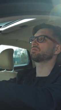 Gözlüklü genç bir adam düşüncelere dalmış, lüks bir arabada rahatça oturuyor, modern bir zarafet yayıyor.