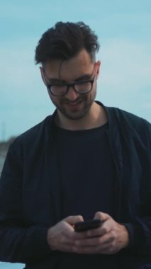 Neşeli genç bir adam mutlu bir şekilde gülümsüyor ve dışarıda güzel bir manzaraya karşı akıllı telefonuyla göz gezdiriyor.