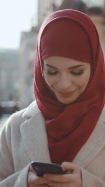 Çarpıcı kırmızı başörtüsü takan şık bir kadın zarif bir şekilde dışarıda akıllı telefonunu kullanırken dikkat çekiyor.