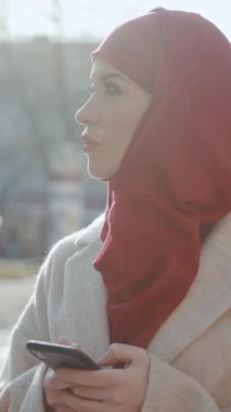 Güzel bir kırmızı başörtüsü takan genç bir kadın akıllı telefonuna derinden odaklanır. Yumuşak, güneşli bir zemin üzerine kuruludur.