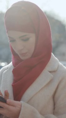 Açık havada parlak kırmızı bir başörtüsü ve krem rengi ceket giyen bir kadının fotoğrafı çekiliyor, dinginlik ve güzellik yayıyor.