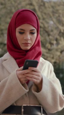Şık bir kırmızı başörtüsü takan genç bir kadın, doğası gereği güzelliklerle çevriliyken akıllı telefonuna odaklanır.