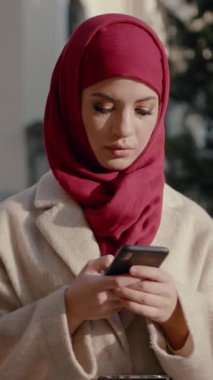 Canlı kırmızı tesettürlü genç bir kadın açık havada otururken akıllı telefonuna derinden odaklanır.