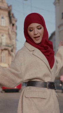 Canlı ve canlı bir şehir ortamında, kırmızı tesettür ve krem rengi ceket giyen genç bir kadın saf zarafet yayar.