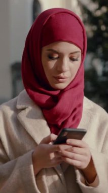 Sakin bir açık hava ortamında şık bir kadın canlı kırmızı başörtüsü takıyor. Akıllı telefonunu kontrol ediyor.