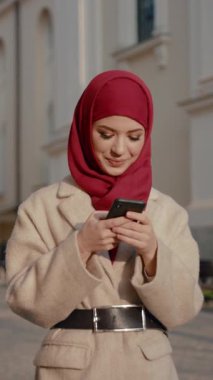 Canlı bir şehir ortamında, kırmızı tesettürlü şık bir kadın günün tadını çıkarırken akıllı telefonunu kontrol eder.