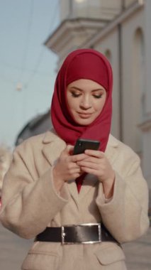 Şık ve şık bir genç kadın canlı kırmızı başörtüsü takıyor. Şehir içinde yürürken akıllı telefonunu kontrol ediyor.