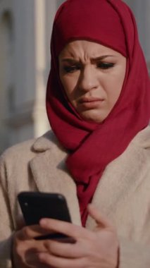 Canlı kırmızı başörtüsü takan bir kadın telefonuna dikkatlice bakarken büyük bir endişe gösterir.