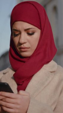 Hareketli bir şehir ortamında, canlı kırmızı başörtüsü takan genç bir kadın akıllı telefonuyla dikkatle iletişim kurar.