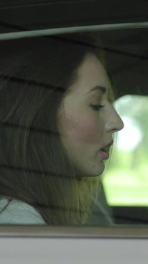 Bir kadın arabanın arkasında oturur, manzara değişirken dikkatlice pencereden dışarı bakar, içsel ruh halini yansıtır.