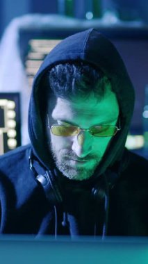 Koyu renk kapüşonlu ve şık güneş gözlüğü takan bir adam gözünün önünde parlak bir bilgisayar ekranına bakıyor.