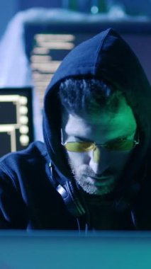 Kapüşonlu ve güneş gözlüklü bir adam dikkatle bilgisayar ekranına bakıyor, görünüşe göre odaklanmış ve görüntülenen içerikle meşgul.