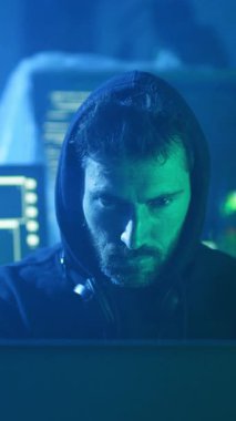 Koyu renk kapüşonlu bir adam dikkatle bilgisayar ekranına bakıyor. Düşüncelere dalmış ya da önemli bir şeye odaklanmış gibi görünüyor.