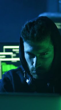 Loş bir odada, kapüşonlu bir adam, dijital bir diyarda gezinirken, gizemli ve odaklanmış bir şekilde dizüstü bilgisayarına bakar.