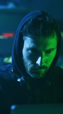 Kapüşonlu bir adam loş ışıklı bir odada oturuyor ve kulaklıklarla dizüstü bilgisayarına odaklanıyor. Sakalı, yaptığı işi yansıtırken kabalık katıyor.