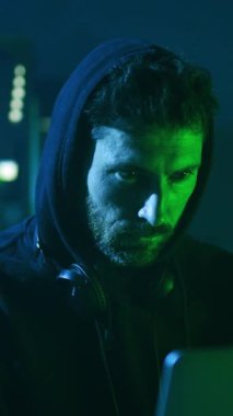 Loş bir odada, kapüşonlu bir adam yoğun bir şekilde bilgisayar ekranına odaklanıyor, teknolojinin cazibesini ve izolasyonunu vurguluyor.