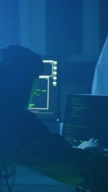 Loş bir odada, kapüşonlu bir figür dizüstü bilgisayara odaklanıyor, yüzü ışıldıyor. Yakınlarda, yeşil bir ekran ürkütücü ortamı güçlendiriyor.