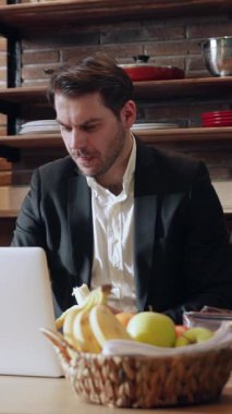 Takım elbiseli bir adam, modern bir çalışma alanında dizüstü bilgisayarıyla ön planda lezzetli meyvelerle çalışır.