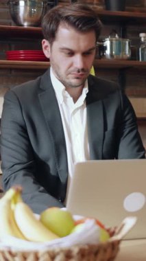 Resmi takım elbiseli bir adam, canlı taze meyvelerle dolu sıcak bir mutfakta dizüstü bilgisayarına odaklanır.