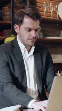 Sivri bir takım elbise giymiş odaklanmış genç bir adam modern çalışma alanında özenle dizüstü bilgisayarı üzerinde çalışıyor.