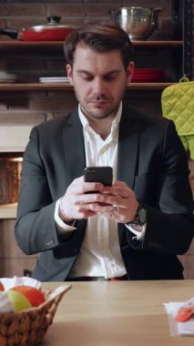 Terzi kıyafeti giymiş profesyonel bir adam şık bir mutfak alanında akıllı telefonuna konsantre oluyor.