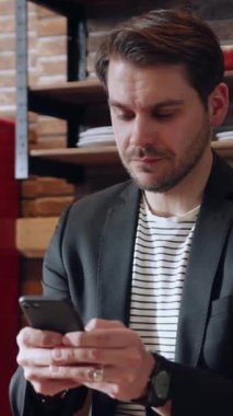 Şık ve modern kıyafetler giymiş bir adam şık bir ofis ortamında otururken akıllı telefonuna odaklanır.