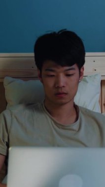 Genç bir adam dikkatle dizüstü bilgisayarı üzerinde çalışıyor, yatakta oturuyor, konsantrasyonu ve nişanı gösteriyor..