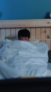 Sakin bir yatak odası ortamında, genç bir adam dikkatli bir şekilde sıcak ve rahat battaniyelerin altından dışarı bakar.