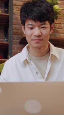 Parlak bir gülümsemesi olan genç bir adam, rahat bir kafede otururken dizüstü bilgisayarıyla coşkuyla etkileşime geçiyor.