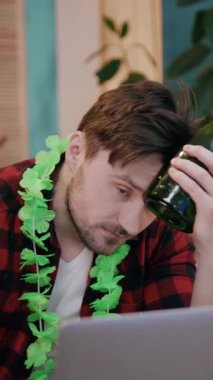 Renkli Hawaii kolyesi takmış hayal kırıklığına uğramış bir adam ev ortamında dizüstü bilgisayarının yanında bira tutuyor.