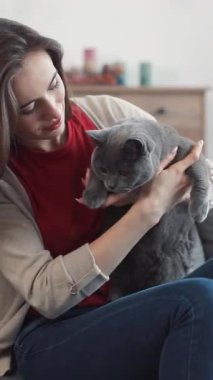 Bir kadın sevgiyle gri bir kediyi kollarında tutuyor ve şık bir kapalı mekan ortamında güçlü bağlarını sergiliyor.