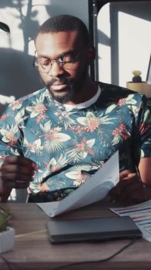 Çiçekli gömlekli bir adam modern bir masada, pencereden gelen sıcak doğal ışıkla aydınlanan belgeler üzerinde çalışıyor.