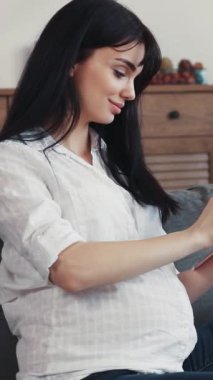 Hamile bir kadın boş zamanlarında evinde rahat ve rahat bir şekilde otururken tabletle sıcak bir şekilde etkileşime girer.