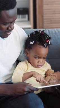 Genç bir çocuk, yumuşak bir oyuncak ayıyı sevgiyle tutarken renkli bir tabletle etkileşime giriyor. Modern ebeveynlik sergiliyor.