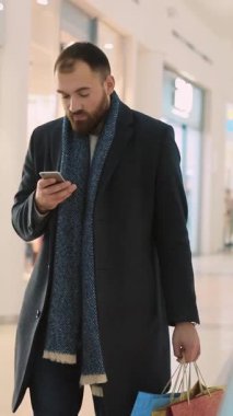 Şık montlu, bakımlı sakallı bir adam alışveriş merkezinde yürürken akıllı telefonunu kontrol ediyor.