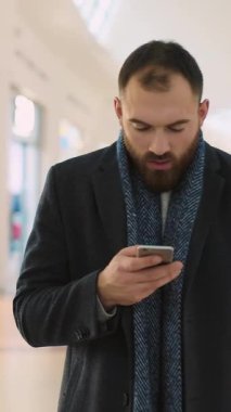 Genç bir adam, yoğun bir alışveriş ortamının koşuşturması ve telaşıyla çevriliyken akıllı telefonuyla meşgul olur.